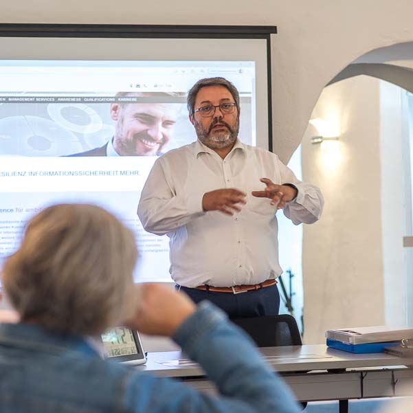 Ein Referent präsentiert vor einem Publikum, während eine Präsentation mit dem Text "RESILIENZ INFORMATIONSSICHERHEIT" auf dem Bildschirm angezeigt wird.