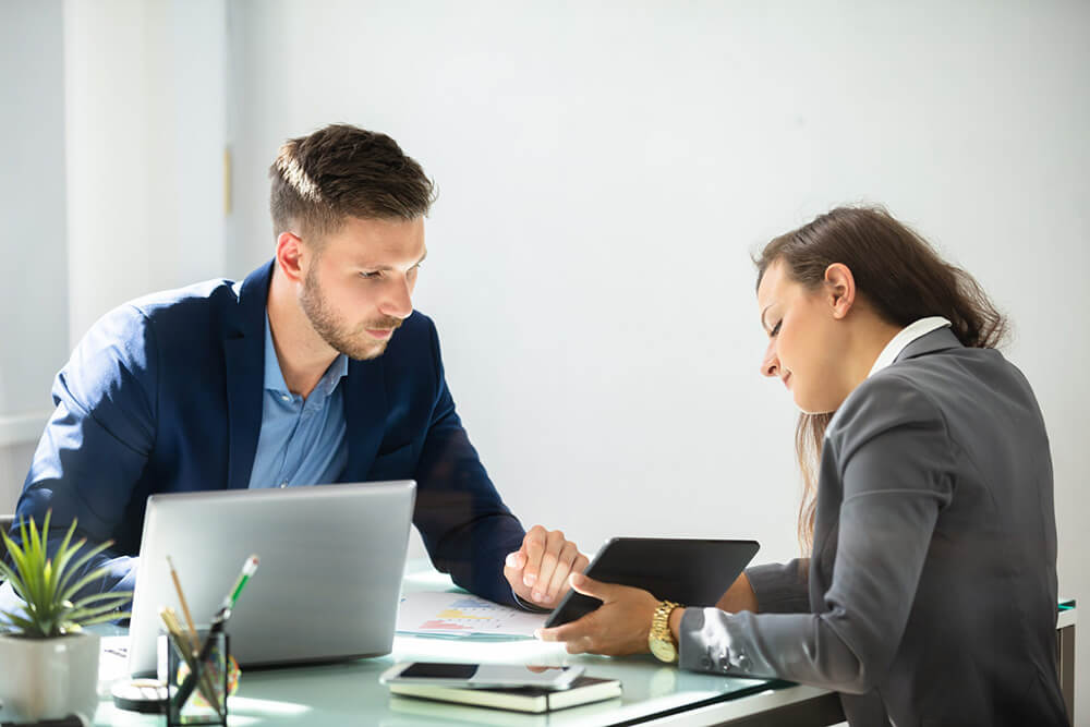 Ein Mann in einem blauen Anzug und eine Frau in einem grauen Blazer sitzen an einem Glastisch in einem hellen Büro. Der Mann schaut auf das Tablet, das die Frau hält, während ein Laptop und Dokumente auf dem Tisch liegen.