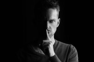 Schwarz-Weiß-Porträt eines Mannes, dessen Gesicht teilweise im Schatten liegt und der einen Finger vertikal über den Mund hält, was ein Geheimnis oder Diskretion symbolisiert. Er blickt direkt in die Kamera.
