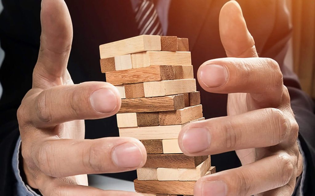 Symbolbild für Risikomanagement oder Geschäftsstrategie: Hände eines Geschäftsmannes versuchen, einen wackeligen Holzturm auf Geschäftsdokumenten vor dem Einsturz zu bewahren.